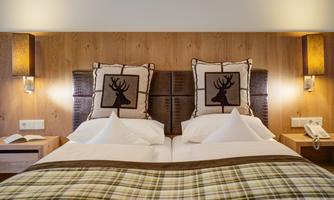 A cozy hotel room with two beds and wooden paneling. The pillows feature a deer motif and the bedding has a checked pattern.