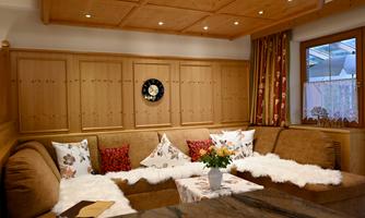 A cozy living room with a large beige couch and colorful cushions. On the table sits a vase with fresh flowers and the room has a warm wooden ceiling.