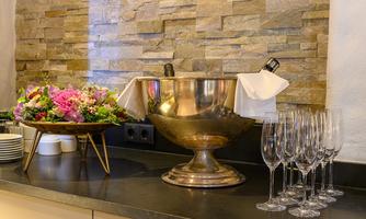 An elegant table with a silver champagne bucket and glass champagne flutes. Next to it is a beautiful bouquet in a golden vessel.