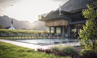 A modern building with a swimming pool and loungers in a quiet setting. In the background, mountains and a church tower are visible.