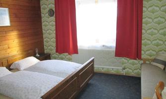 A simple bedroom with two beds and a sofa. The walls are decorated with a green pattern and wooden paneling, and the windows have red curtains.