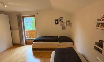 A bright bedroom with two single beds and a large window. The floor is made of wood and a round carpet lies on the floor.