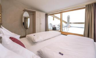 A modern bedroom with a large window and a view of a balcony. The decor is simple and elegant, featuring a large bed and a mirror.