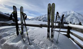 Skis are standing in the snow next to a wooden railing. In the background, there are snow-covered mountains and a blue sky.