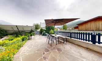 A beautiful outdoor area with a table and chairs under a sunshade. Surrounded by colorful flowers and a green landscape.