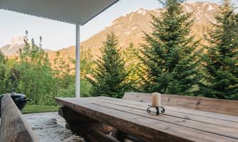 A wooden log table is set outdoors, surrounded by tall fir trees and a picturesque mountain landscape. On the table, there is a candle that creates a cozy atmosphere.