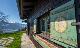 A charming wooden cottage with colorful shutters and a view of the Alps. The green meadow and the mountains in the background create an idyllic atmosphere.