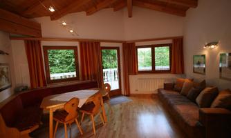 A cozy living room with a wooden ceiling and large windows. There is a dining table and a couch, surrounded by warm colors and a view of the greenery.