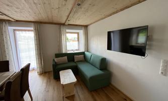 A cozy living room with a green sofa and a flat-screen TV. Large windows provide plenty of daylight.