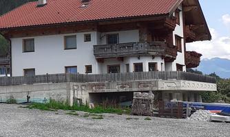 A traditional Alpine house with a red tiled roof and balconies. Surrounded by a gravel area and mountains in the background.