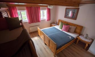 A cozy bedroom with wooden furniture and bright shutters. There is a double bed and an additional sleeping place on the left side.