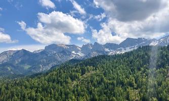 A picturesque mountain landscape with snow-capped peaks and a forest full of green trees. The sky is blue with some white clouds.