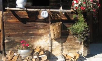A rustic wooden wall with a basket, flowers, and a small collection of firewood. Above the basket, some flowers are hanging in flower pots.