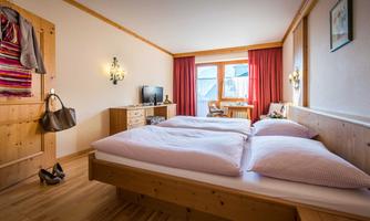 A cozy bedroom with two beds and wooden decor. There is a television, a chair, and large windows with red curtains that let in plenty of light.