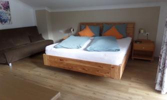 A cozy bedroom with a large double bed and orange cushions. Next to the bed, there is a sofa and a bedside table.