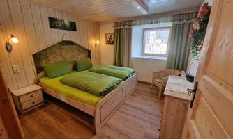 A cozy room with two green beds and warm wood paneling. Windows with curtains and a small seating area enhance the homely atmosphere.