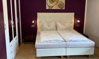 A cozy bedroom with a white bed and purple walls. In the background, there is a wardrobe and a decorative picture.