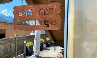 A decorative sign with the inscription "Grill & Chill Lounge". In the background, a well-kept garden and sunny weather conditions can be seen.