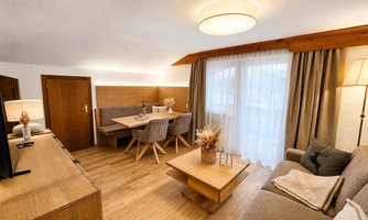 A modern living room with a cozy sofa, a wooden table, and a dining area. Large windows let in plenty of light and the decor is warm and inviting.