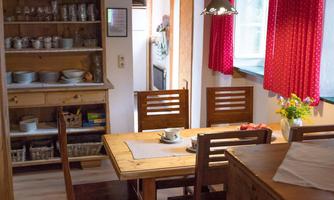 A cozy kitchen with a wooden table and chairs. Red curtains and dishes on the shelf give the room a warm atmosphere.