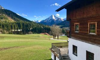 A picturesque landscape with a wooden house and snow-covered mountains in the background. The meadows are green and the sky is clear and blue.