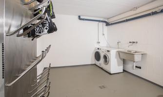 A clean laundry room with a washing machine, a sink, and a shelf for clothes. The walls are white and the floor is gray.