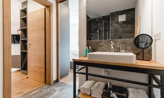 A modern bathroom with an elegant wash station and wooden shelf. The walls are in dark stone and there is a large shower.