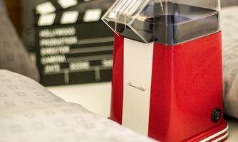 A red popcorn maker is on a bed, surrounded by pillows. In the background, there is a clapperboard.