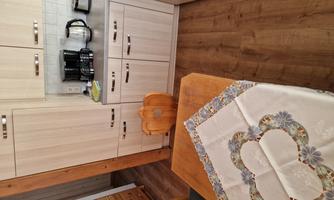 A bright kitchen with a wooden table and a tablecloth. In the background, kitchen cabinets and a coffee machine can be seen.