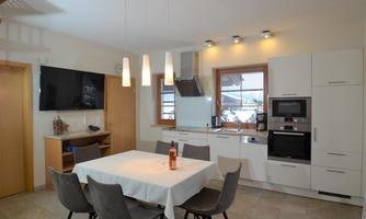A modern kitchen with a large dining area. The table is covered with a white tablecloth and there is a television on the wall.