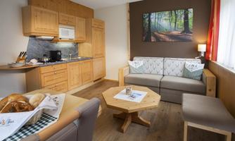 A cozy living room with a modern kitchen and a wooden table. A picture of a forest hangs on the wall, creating a tranquil atmosphere.