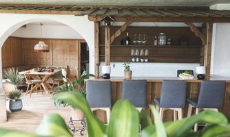 A cozy interior view with a wooden bar and chairs. In the background, a dining table and plants can be seen.