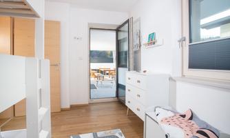 A bright children's room with a bunk bed, a dresser, and a carpet. The open door leads to a balcony or terrace.