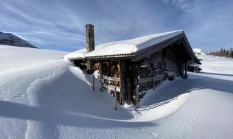 A snow-covered cabin stands in the midst of a white landscape. The sky is clear and blue.