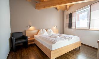 A modern bedroom with a large bed and two cushions. There is a comfortable chair and bright wood interior.