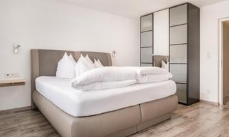 A modern bedroom with a large bed and elegant pillows. The room is bright and friendly, with a wardrobe next to the bed.
