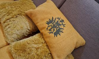 An elongated cushion with an eagle design lies on a gray couch. Surrounding the cushion are several yellow, fluffy cushions.