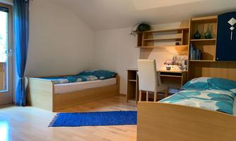 A bright room with two single beds and a desk. The room has hardwood flooring and large windows with curtains.