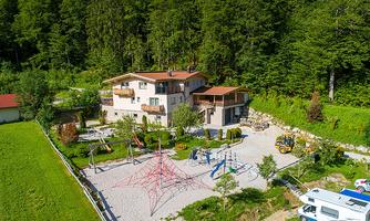 A beautiful building surrounded by trees, with a playground in the foreground. Various play equipment and a slide can be seen in the area.