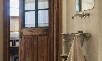 A rustic entrance area with a wooden door and a modern sink. A towel hangs on a chair, surrounded by natural stones.