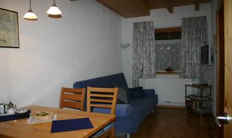 A cozy living room with a blue sofa and a wooden table. Curtains hang in front of the window, and there are some pieces of furniture and accessories visible.