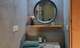 A modern bathroom with a round mirror and a wooden shelf. On the shelf, there is a vase with flowers and personal care products.
