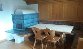 A cozy dining area with a large table and wooden chairs. In the background, there is a nostalgic blue stove and wooden-paneled walls.