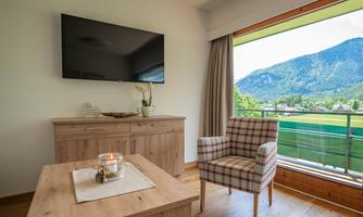 A modernly furnished room with a television, a wooden table, and a cozy chair. Large windows provide a view of the mountains and the green landscape.