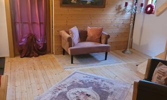 A cozy living space with wooden flooring and a comfortable armchair. A picture hangs on the wall, and a beautiful rug lies on the floor.