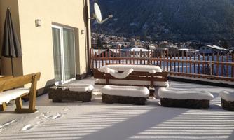 A terrace with snow and stone seating. In the background, mountains and houses can be seen.