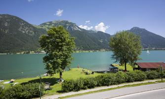 A quiet lake surrounded by green meadows and mountains. In the background, there are trees and a small wooden house.