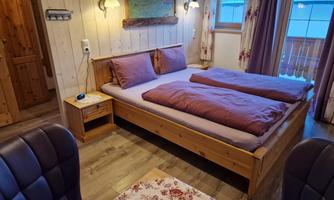A cozy bedroom with a wooden bed and purple bedding. The room has a small table and windows overlooking the outdoors.