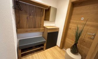 A bright hallway with a wooden shelf, a bench, and a small refrigerator. A coat rack hangs on the wall, and a plant stands in the corner.