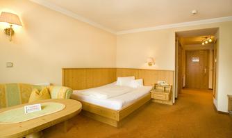 A simple room with a bed, a sofa, and a table. The walls are light and made of wood, and there is a warm atmosphere.